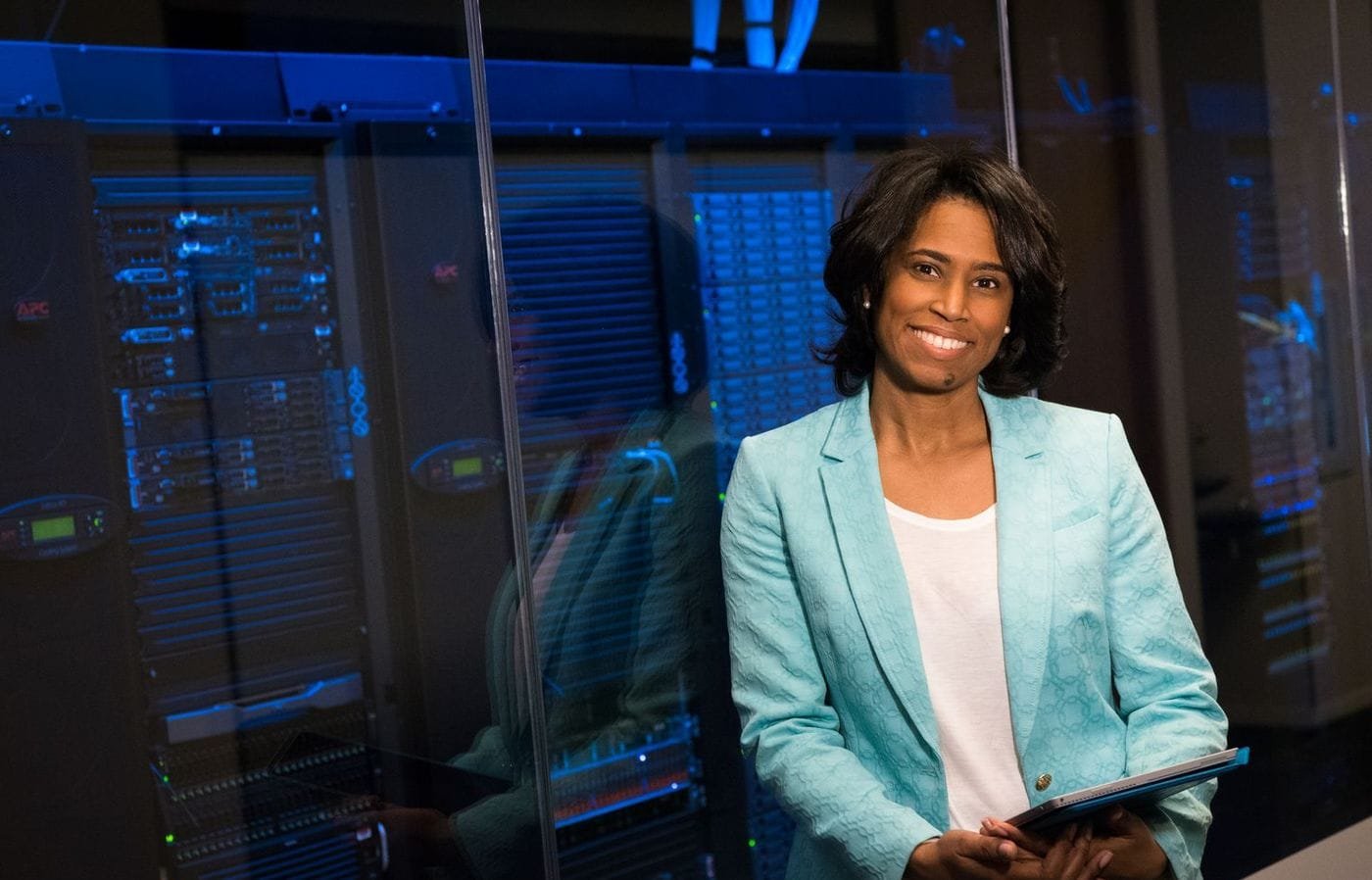 A smiling IT professional in a teal blazer holds a tablet beside glass-fronted server racks in a data centre.
