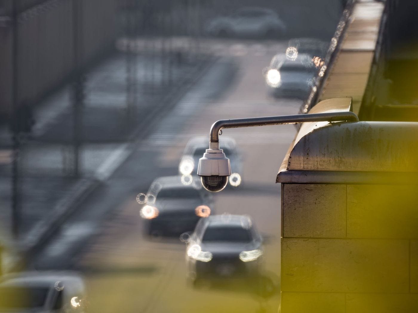 A dome CCTV camera on a wall-mounted arm overlooks a busy road with blurred car headlights.