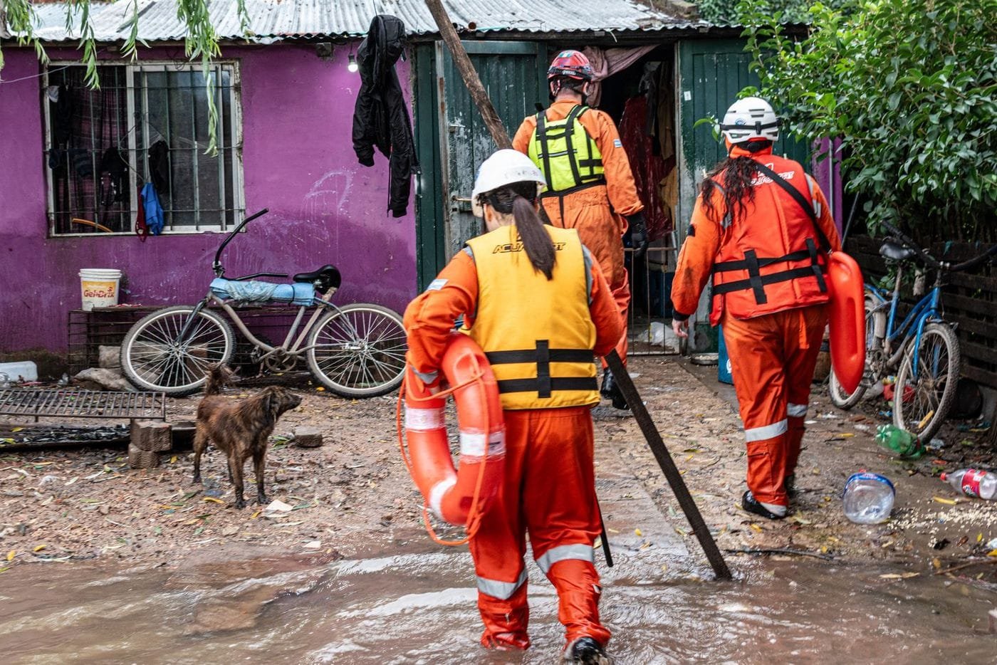 Rescue workers in orange suits, helmets and life vests wade through a flooded, muddy street carrying a lifebuoy.