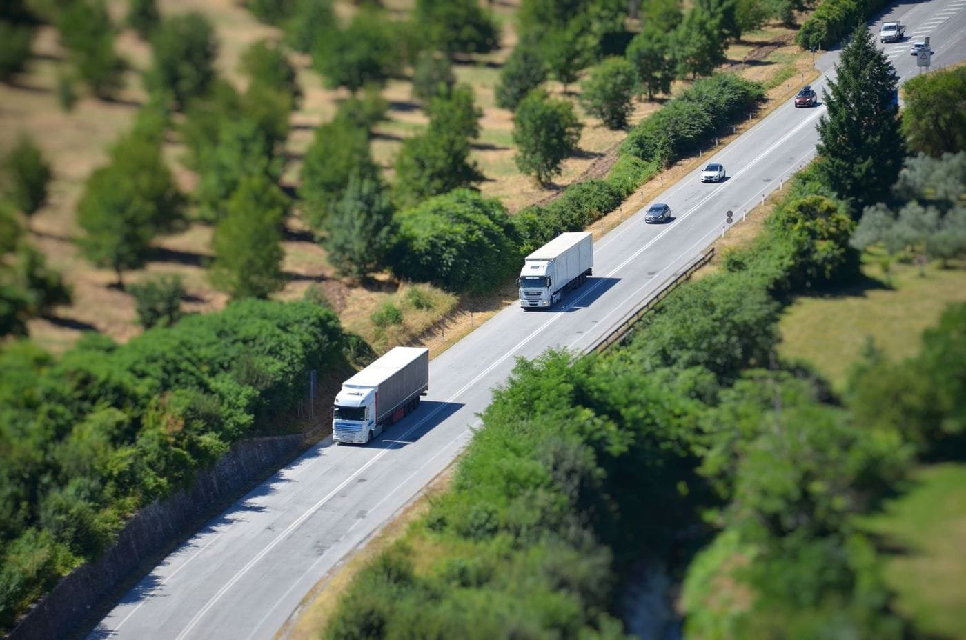 Aerial view of two white freight trucks and several cars driving along a highway through green countryside.