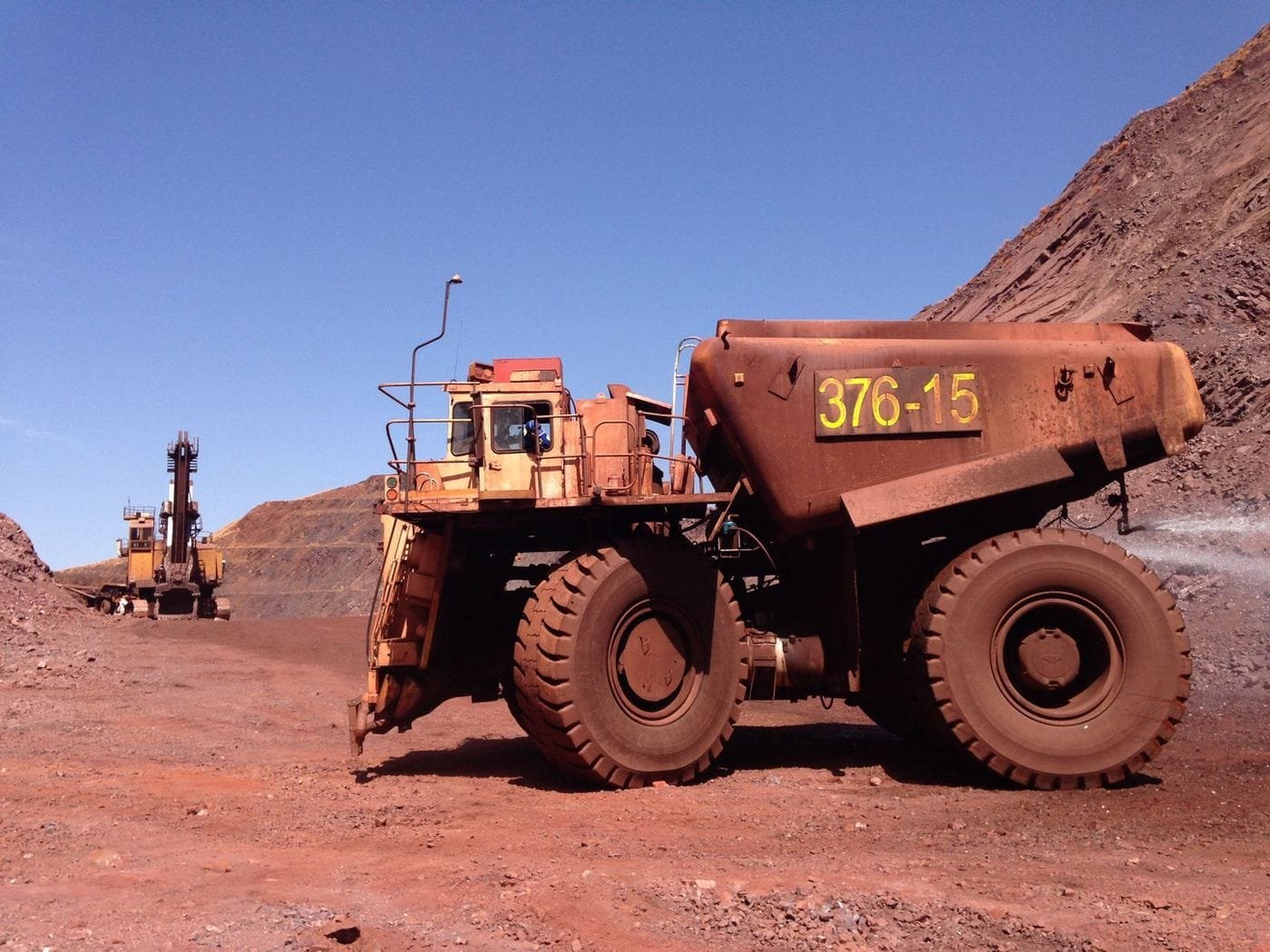 A large haul truck and an excavator stand on the red earth of an open-pit mine under a clear blue sky.