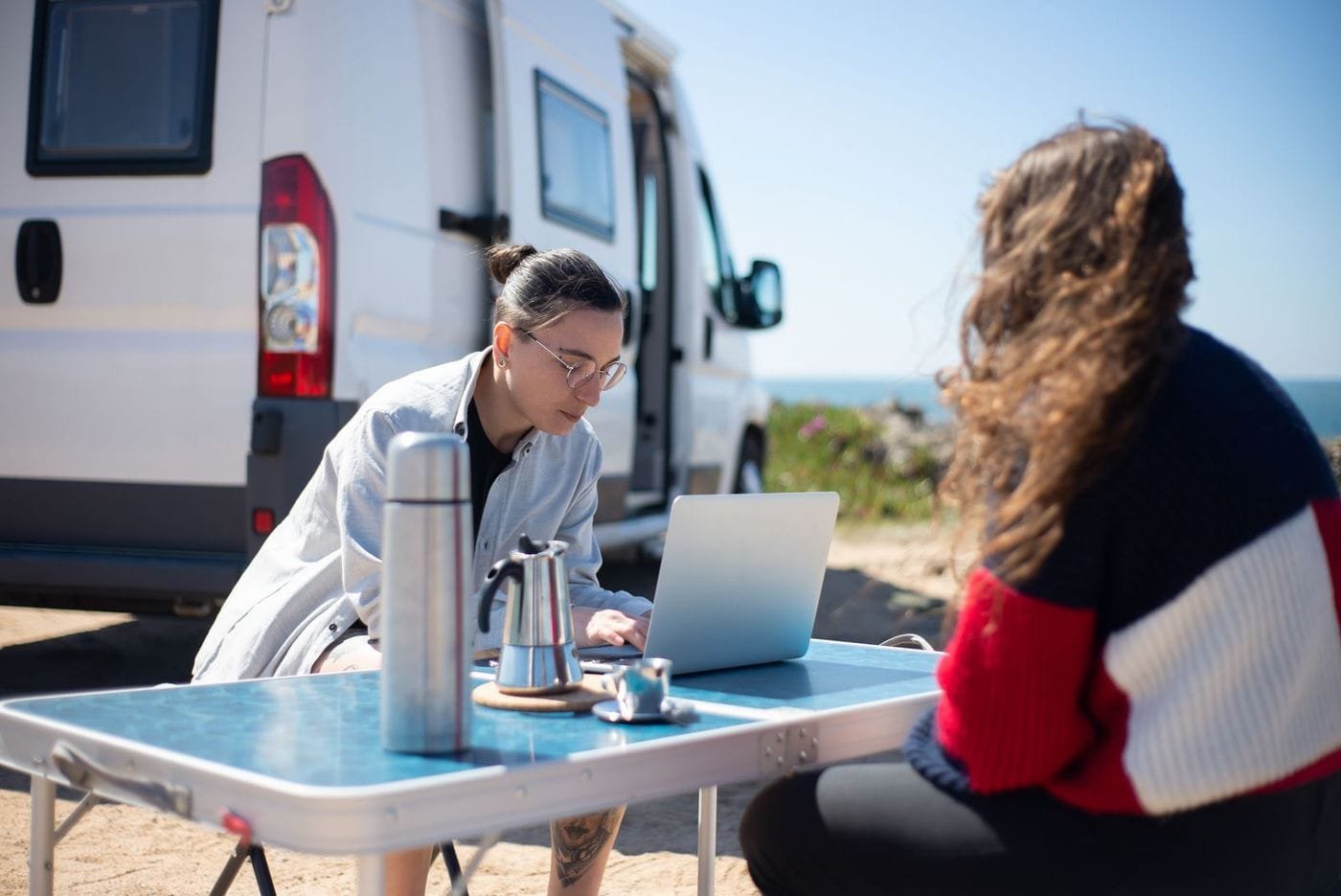 A person works on a laptop at a folding table beside a white camper van at a sunny seaside spot while a companion looks out to sea.