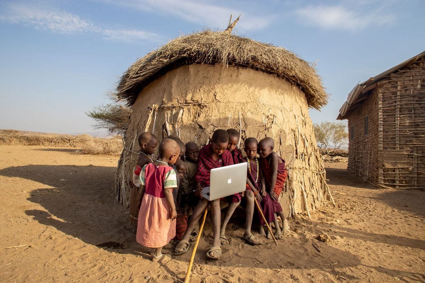 African children gather around an open laptop in front of a thatched mud hut in a dry rural landscape.