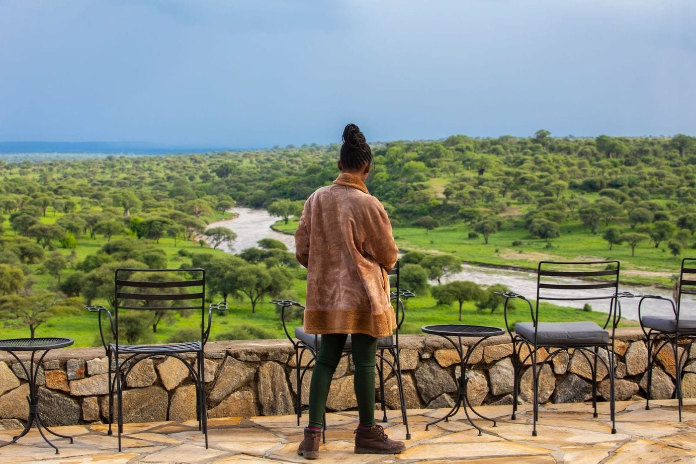 A guest stands on a stone terrace with cafe tables and chairs overlooking a green river valley and bush woodland.