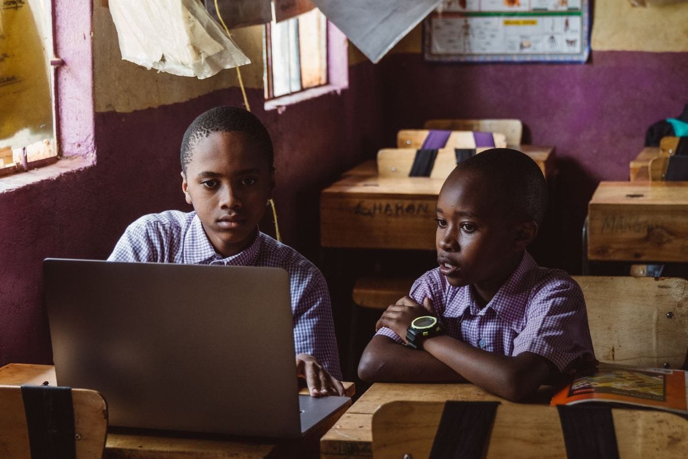 Two schoolboys in uniform shirts share a laptop at wooden desks in a classroom.