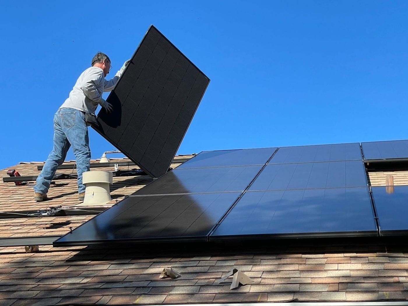 A worker lifts a large black solar panel into place on a tiled roof under a clear blue sky.