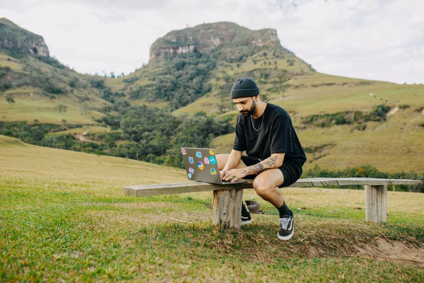 A man in a beanie sits on a wooden bench working on a laptop in green grassy hills with a rocky peak behind him.